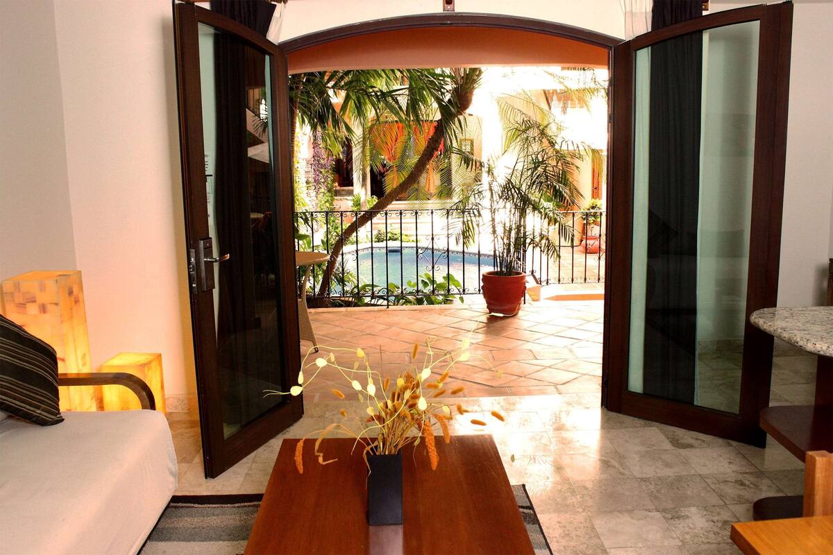 Una habitación con una cama, una mesa de madera con un jarrón de plantas secas y vistas a una piscina y zona exterior con plantas y una valla.