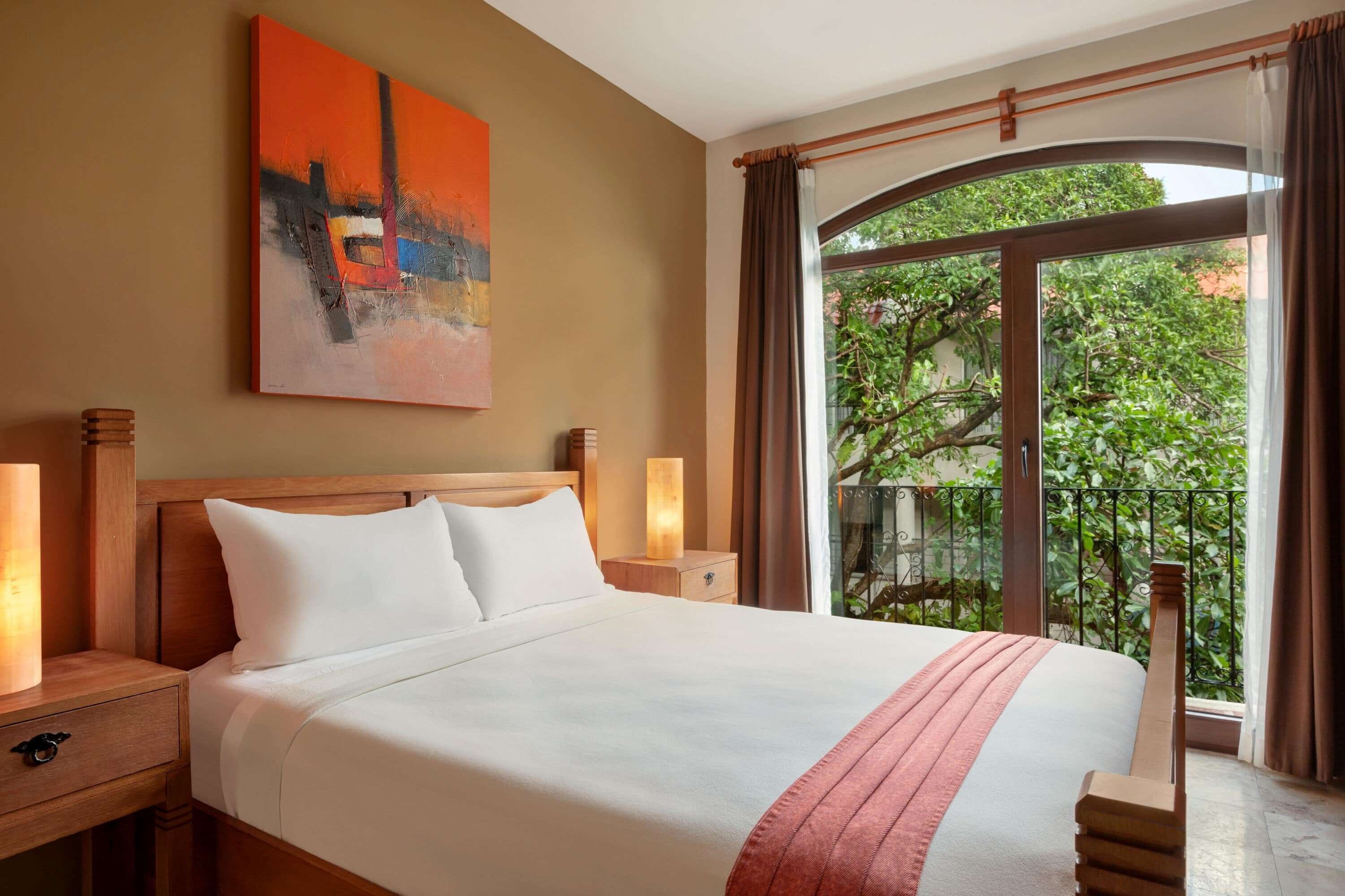 Une chambre d’hôtel avec un grand lit, une table de chevet, une lampe, un tableau au mur et un balcon donnant sur un paysage verdoyant.