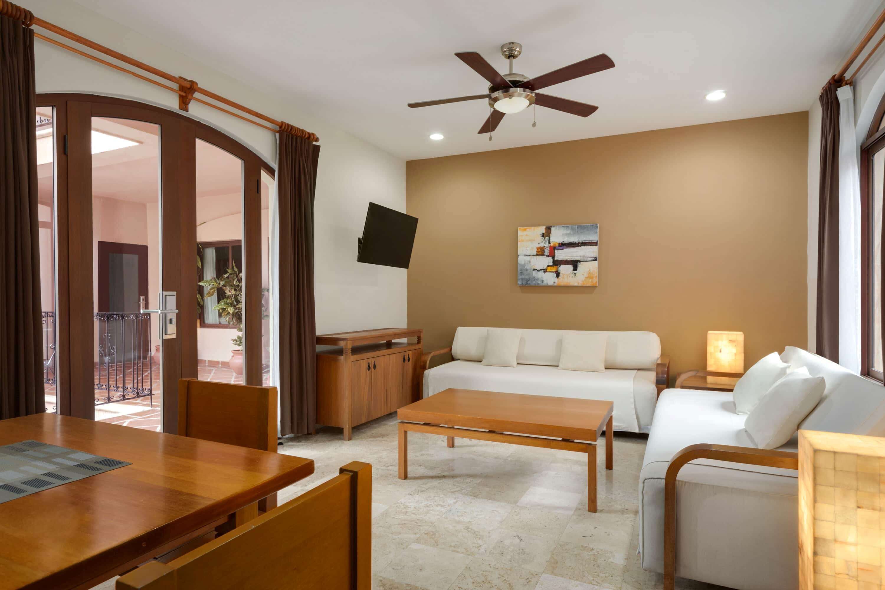 A modern living room with a sofa, coffee table, and wooden furniture. A ceiling fan and TV are mounted on the wall.