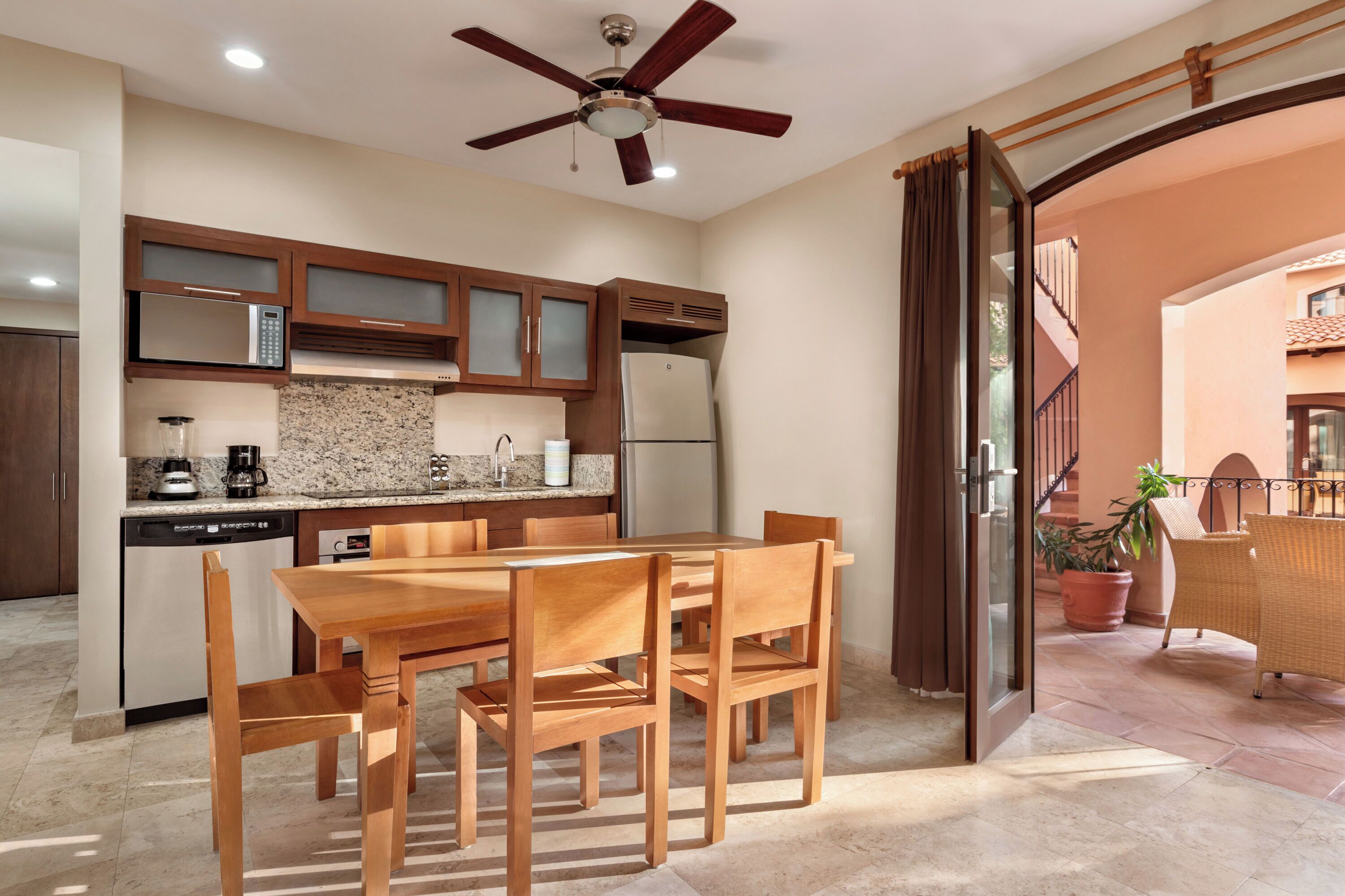 A modern kitchen with wooden cabinets, a built-in microwave, and a refrigerator. There is a dining table with chairs and a ceiling fan.