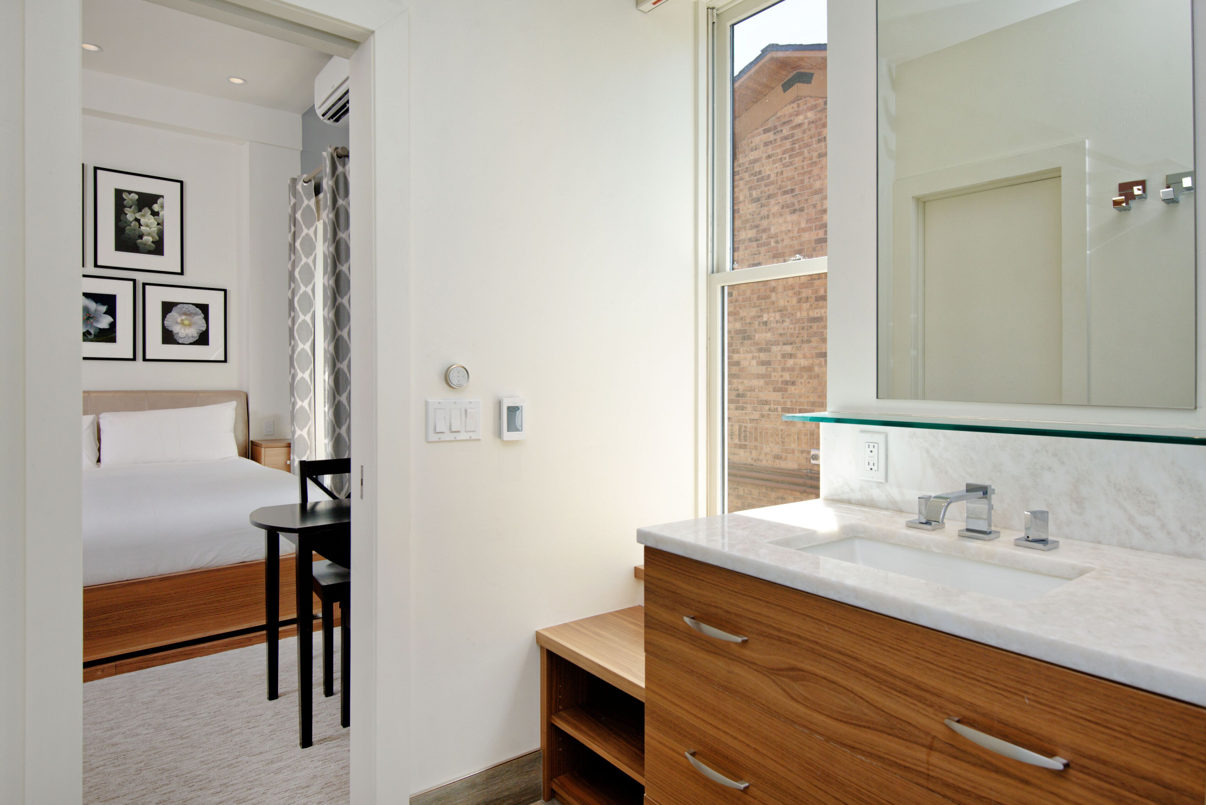 A hotel room with a bed, bedside table, chair, wooden cabinet, and a bathroom with a sink and mirror.