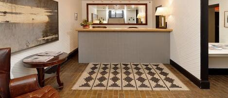 Zona de recepción de hotel con un espejo grande, un mostrador de recepción, una alfombra estampada, un sillón de cuero marrón y una obra de arte colgada en la pared.