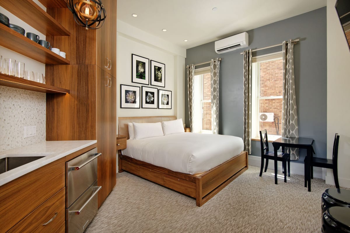 A bedroom with a wooden bed, a desk, and a chair. There are shelves with dishes and glasses, and an air conditioning unit on the wall.
