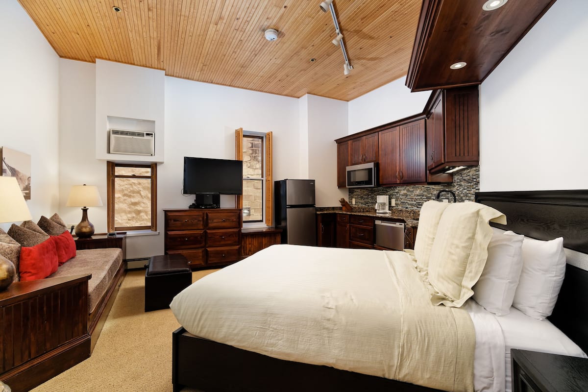 A bedroom with a large bed, a TV, a dresser, a desk, and a kitchenette.