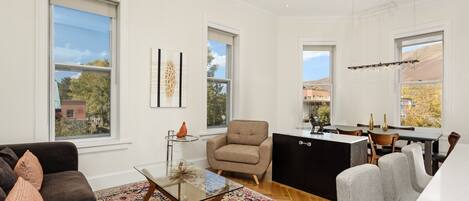 A spacious living room with a sofa, a glass coffee table, and a patterned rug. There is a beige armchair and a dining area with a table and chairs. Large windows provide views of the outside.