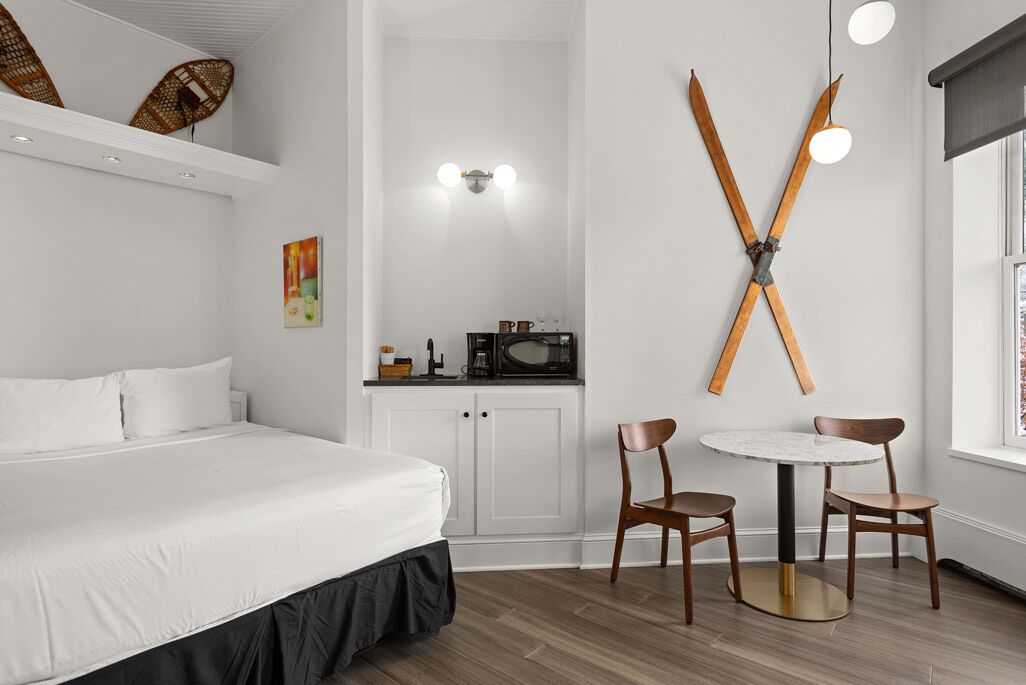 A hotel room with a bed, a small kitchenette, a dining area with a table and chairs, and a wall-mounted ski rack.