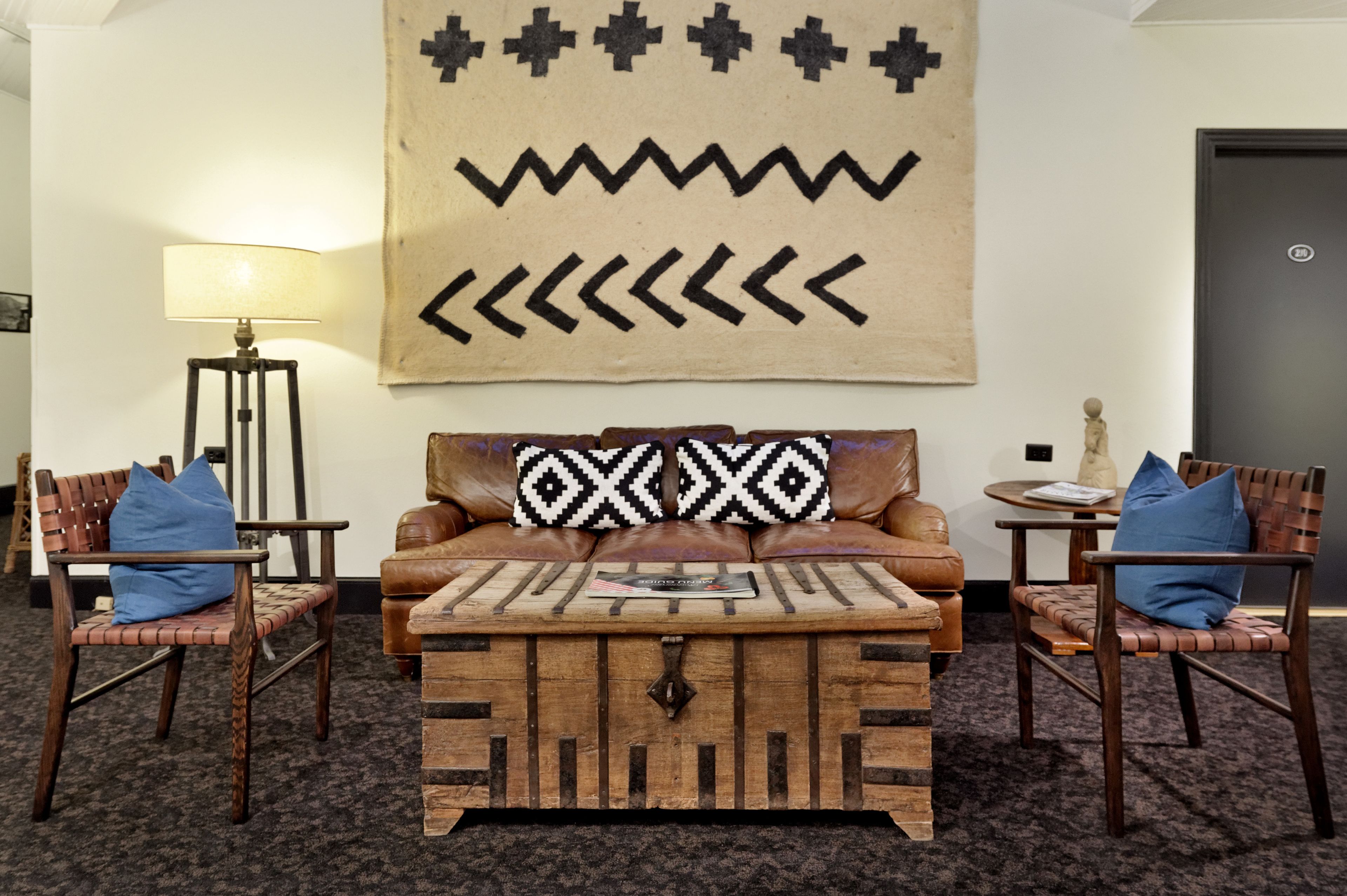 A room with a brown leather sofa, two wooden chairs, a small wooden coffee table, and a wall hanging with geometric patterns.
