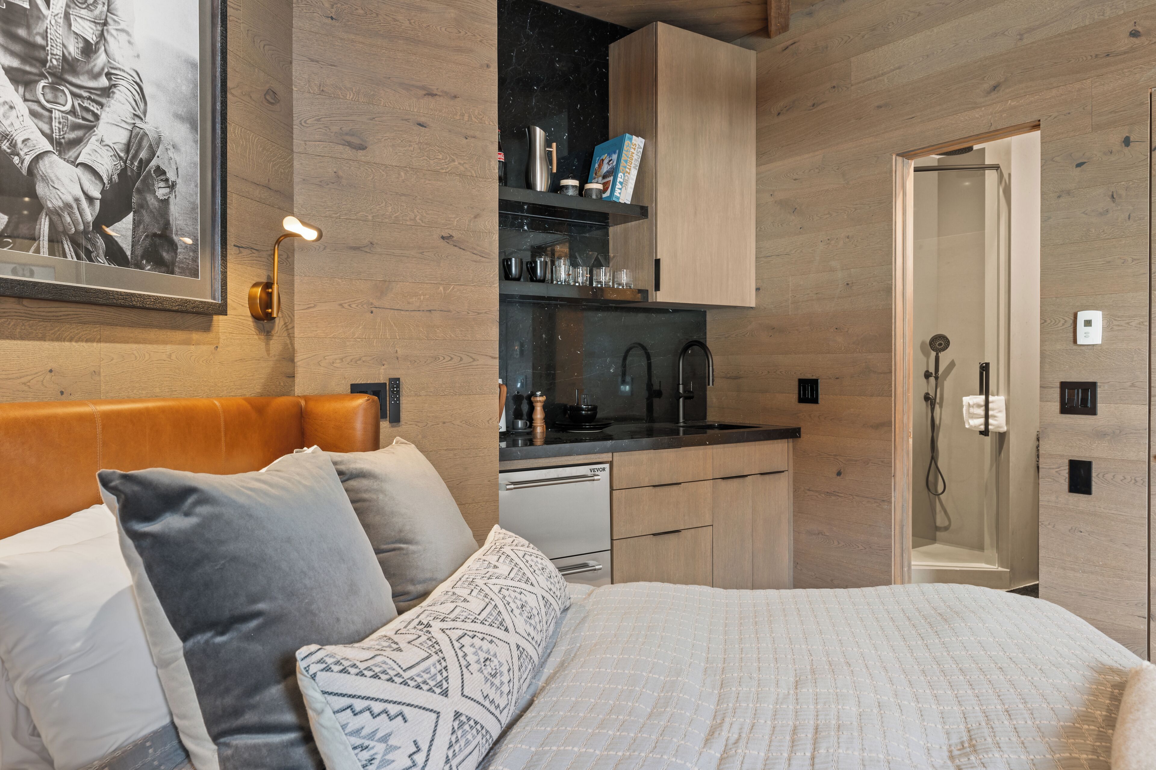 Une chambre à coucher comprenant un lit, des oreillers, une table de chevet, une tête de lit en bois, un tableau encadré au mur, une cuisine intégrée avec un évier et des placards, et un dressing.