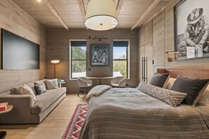 A modern bedroom with a large bed, a sofa, a desk, and a chair. There is a painting on the wall and a window with a view of the outdoors.