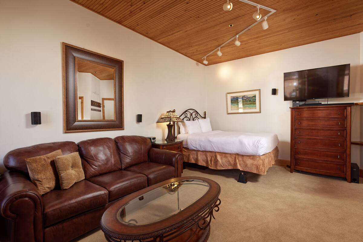 A bedroom with a wooden ceiling, a brown leather sofa, a glass coffee table, a bed with white bedding, a dresser, and a flat-screen TV.