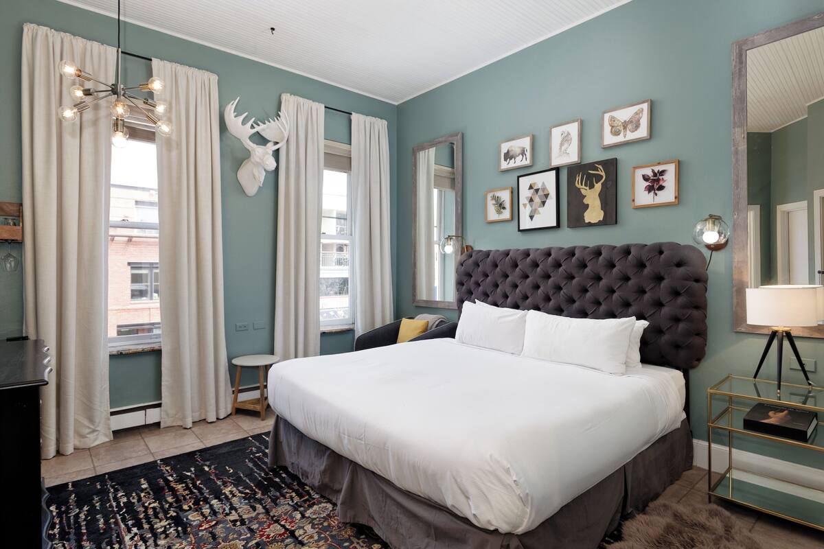 A bedroom with a large bed, a tufted headboard, a window with curtains, and wall art.