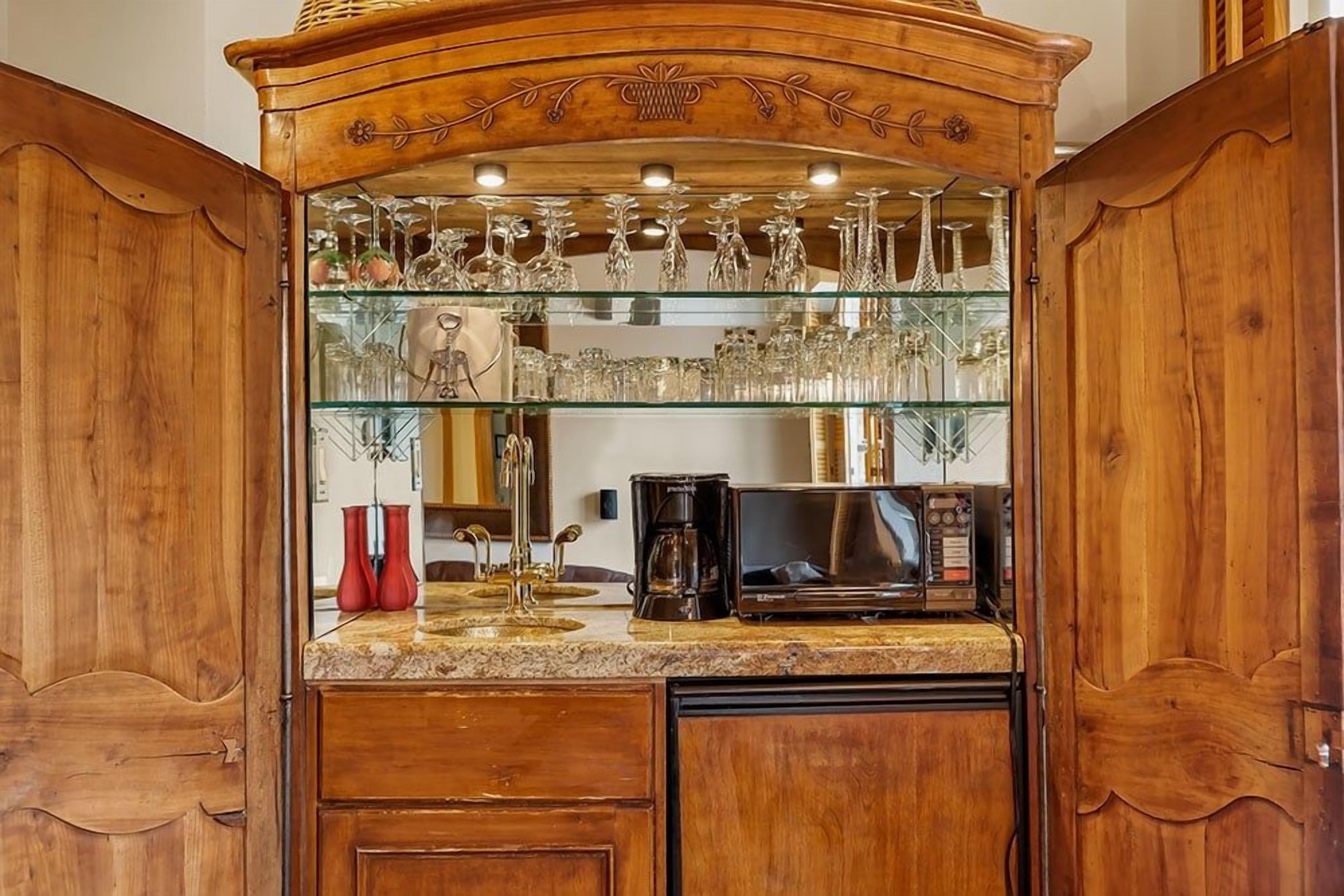 A wooden bar area with glass shelves displaying various glassware, a coffee maker, and a microwave.