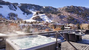 Un bain à remous sur une terrasse en bois, avec vue sur une montagne enneigée et des immeubles résidentiels.