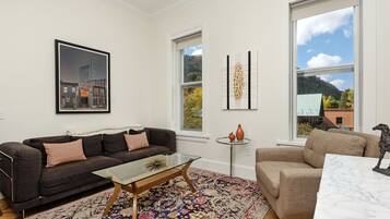 Ein Wohnzimmer mit einem dunkelbraunen Sofa, einem Glastisch, einem beige Armlehnstuhl und einem weißen Marmortisch. Zwei Fenster bieten Blick auf einen Berg und ein Gebäude.