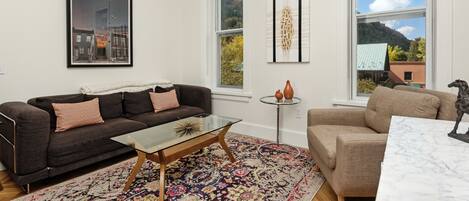 A living room with a dark brown sofa, a glass coffee table, a beige armchair, and a white marble table. There are two windows with views of a mountain and a building.