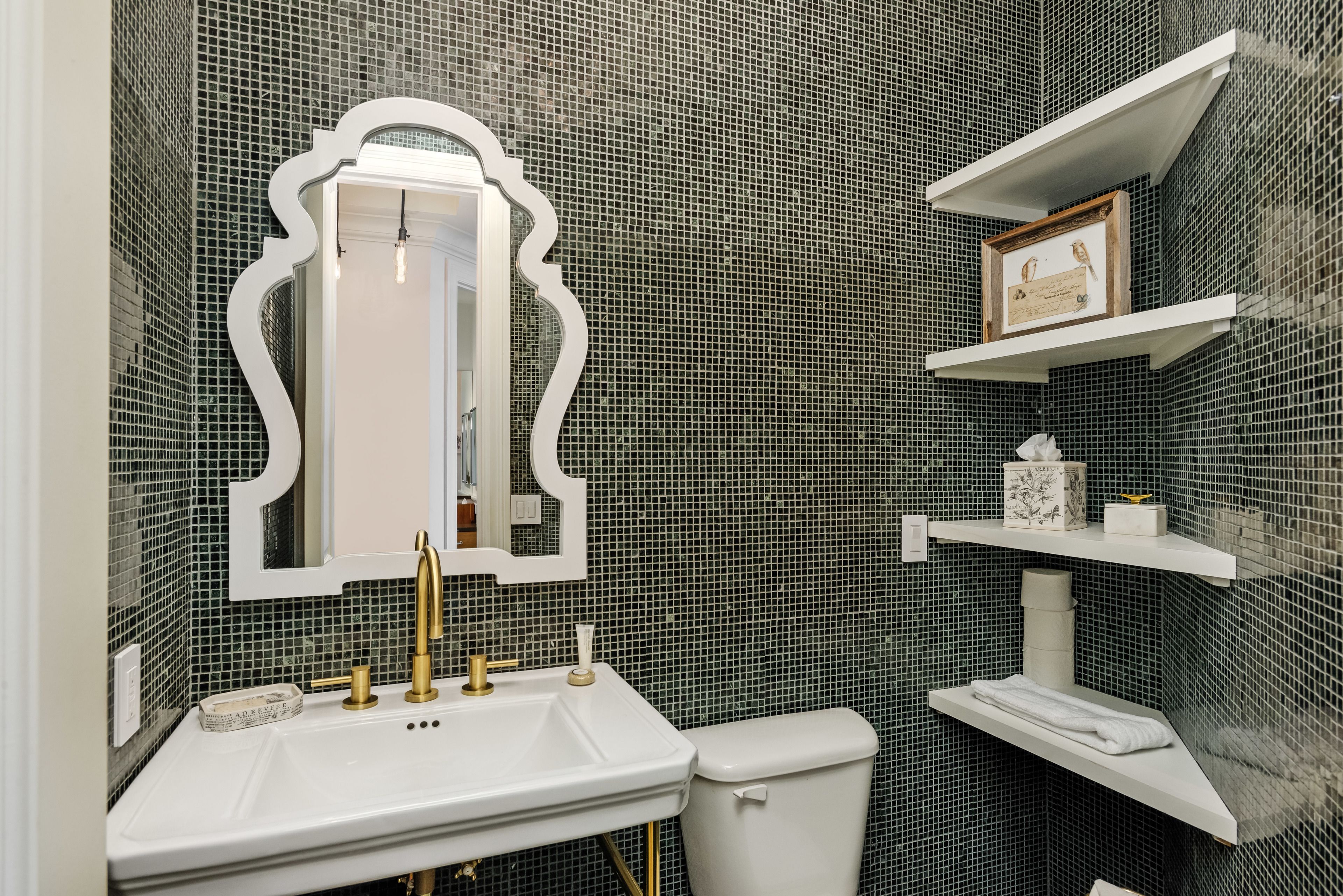 Un bagno con lavandino bianco, rubinetti dorati, un water bianco e uno specchio con cornice decorativa. Si notano mensole con articoli da toilette e un quadro appeso alla parete.