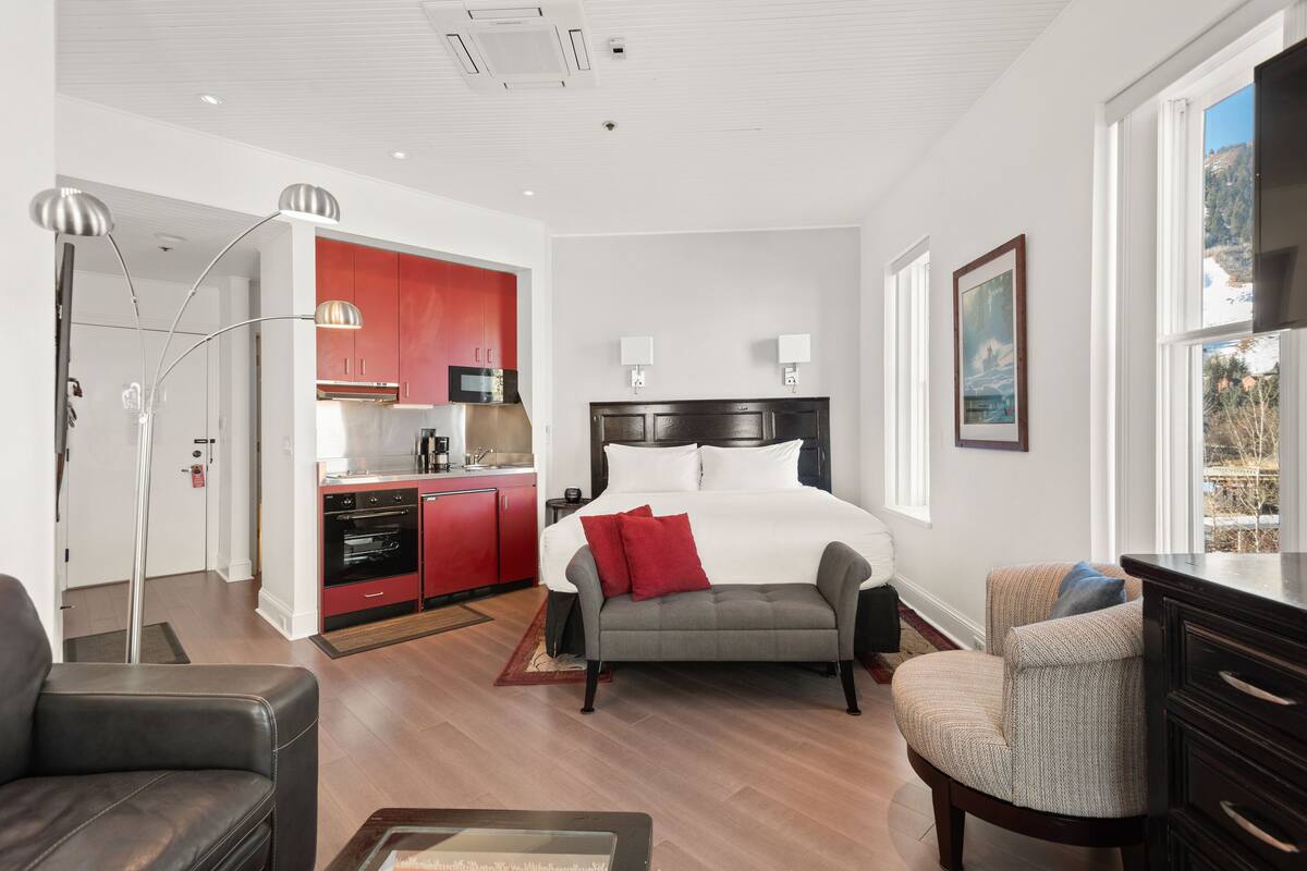 A modern hotel room with a large bed, a sofa, a dining area with red cabinets, and a view of the outdoors.