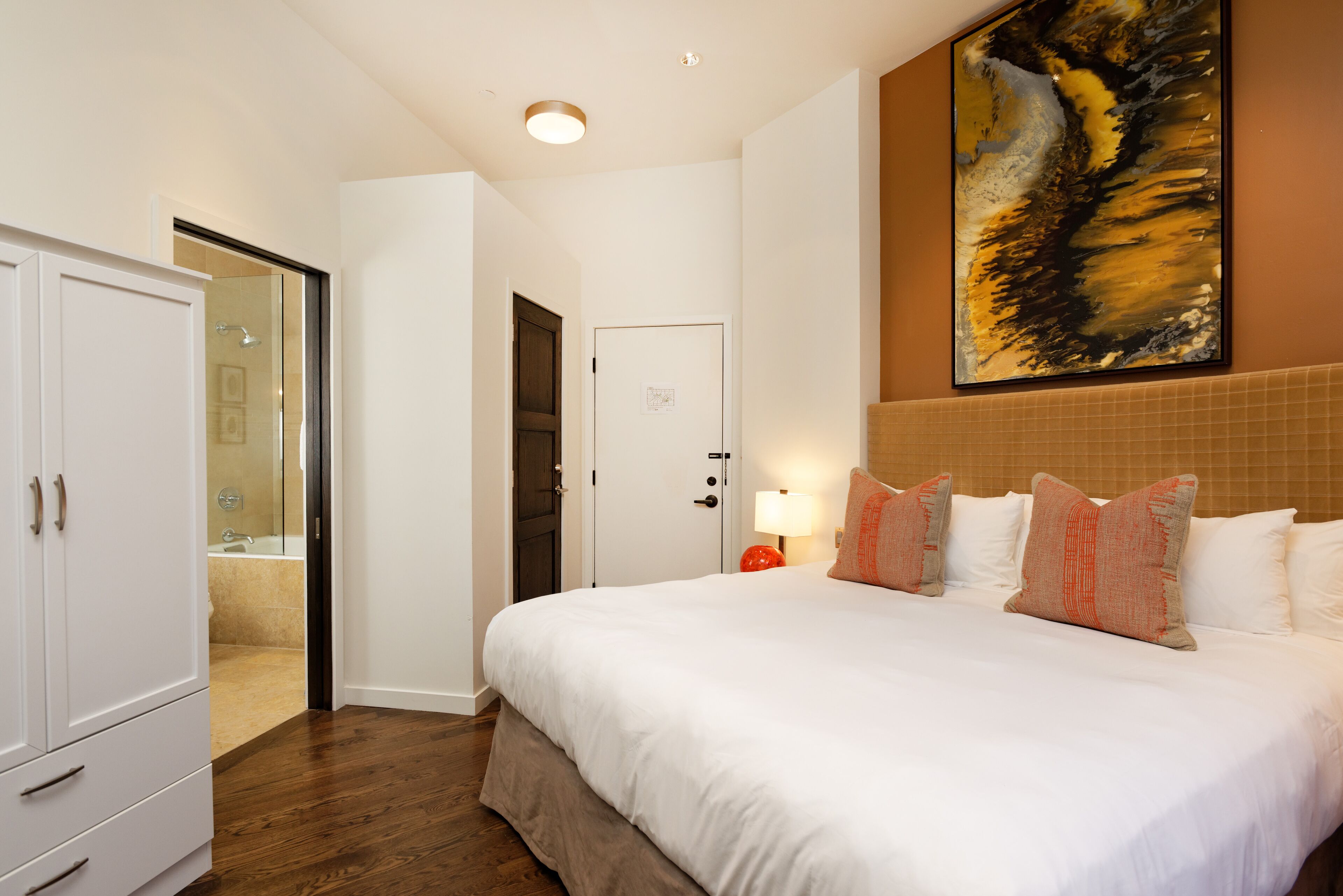 Habitación de hotel con una cama grande, un armario, un baño con puerta de cristal y un cuadro en la pared.