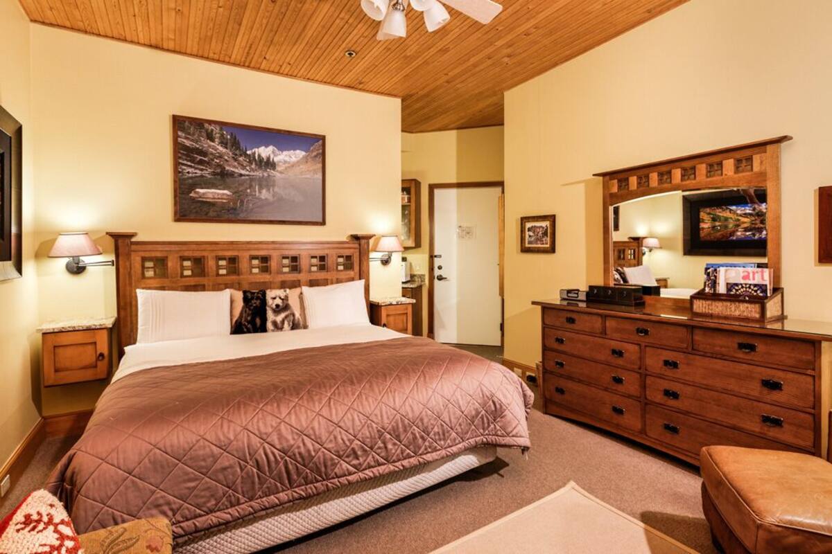 A bedroom with a large bed, wooden headboard, a painting of a mountain landscape, a dresser, and a television.
