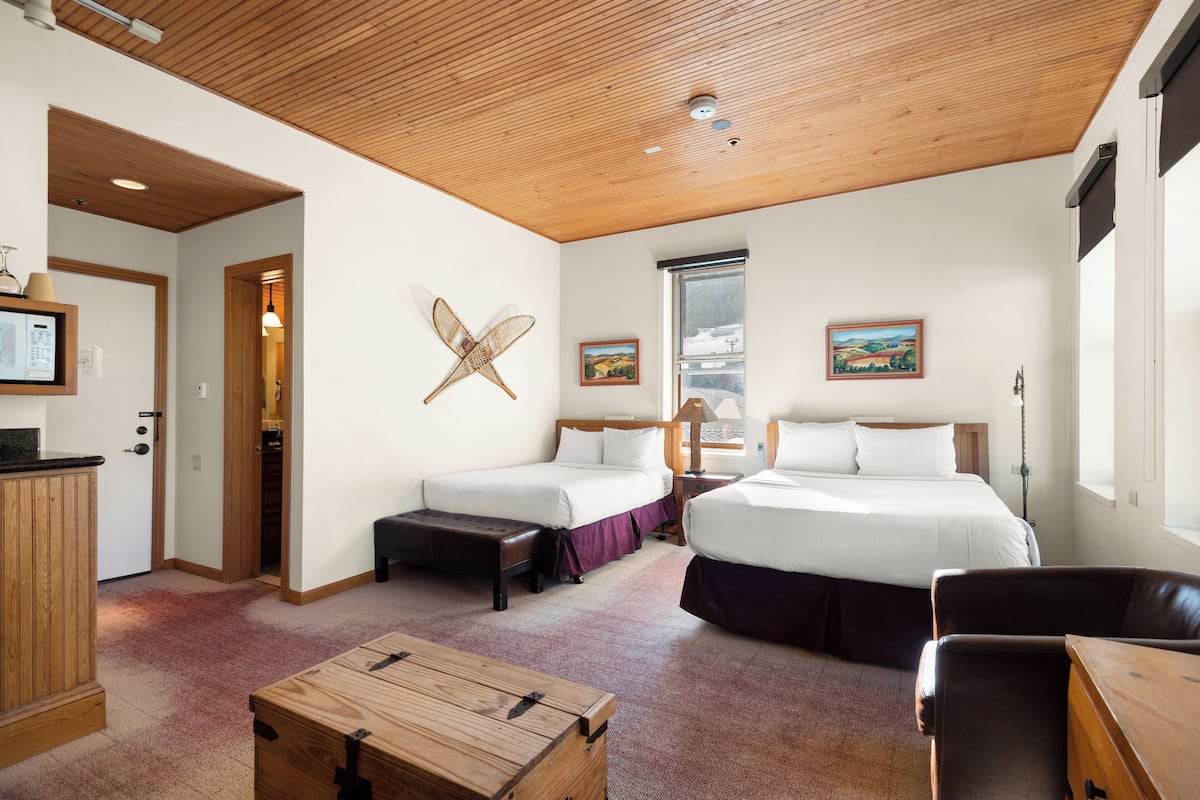 A hotel room with two beds, a wooden ceiling, a desk, and a window with a view.