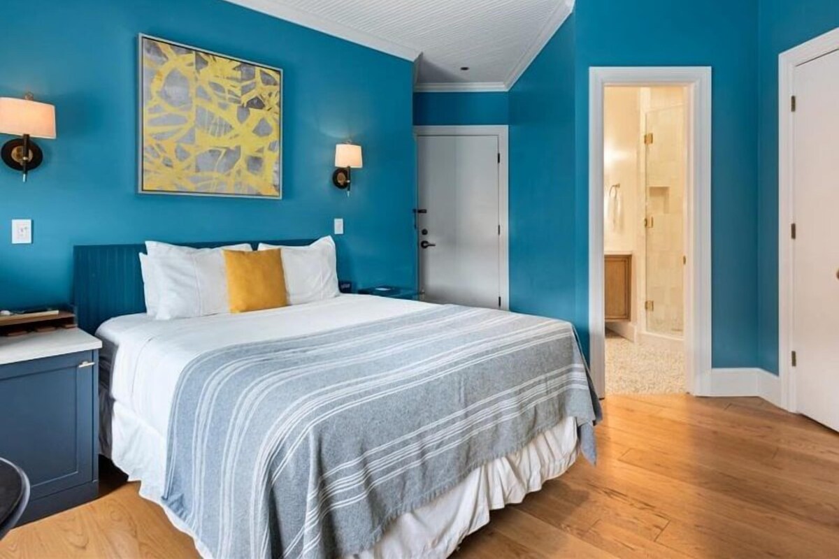 A bedroom with a bed, a nightstand, a lamp, a painting, and a door leading to another room.