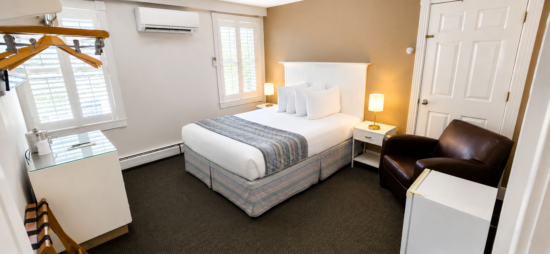 A hotel room with a bed, bedside tables, a chair, a wardrobe, and an air conditioning unit.