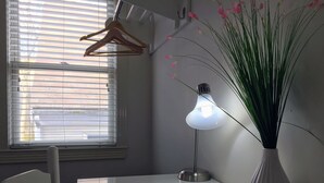 A room with a white table, a hanging light fixture, a white vase with green plants, and a window with blinds.