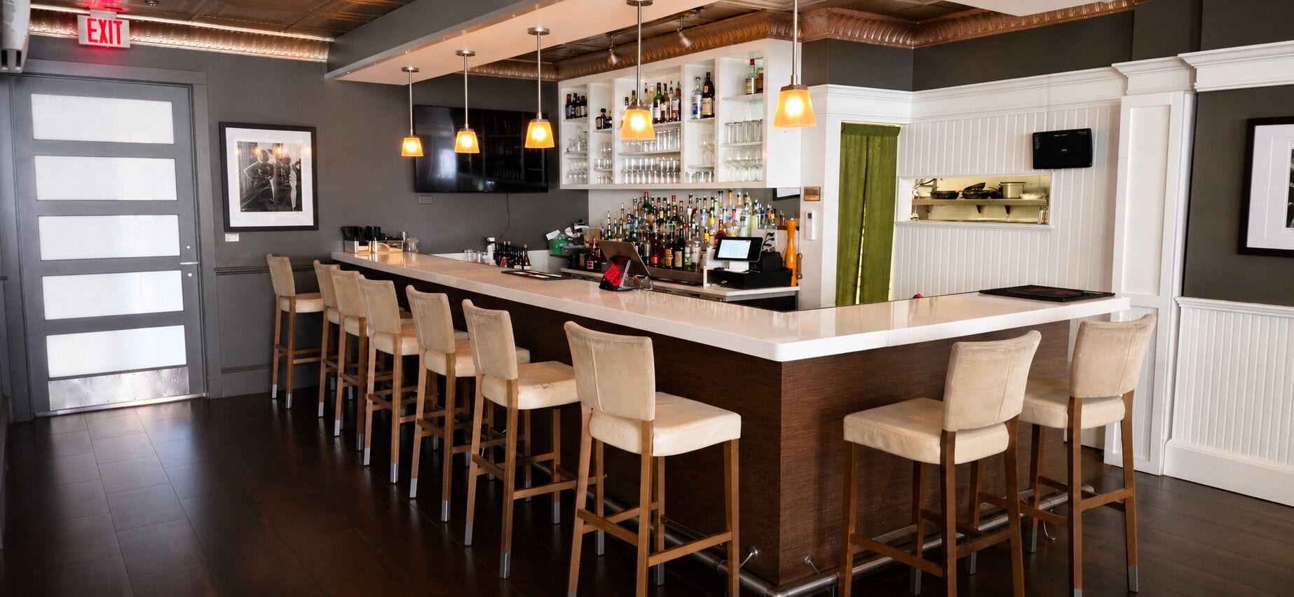 A bar area with a long counter, multiple stools, a television mounted above the bar, and a variety of bottles and glasses on shelves.