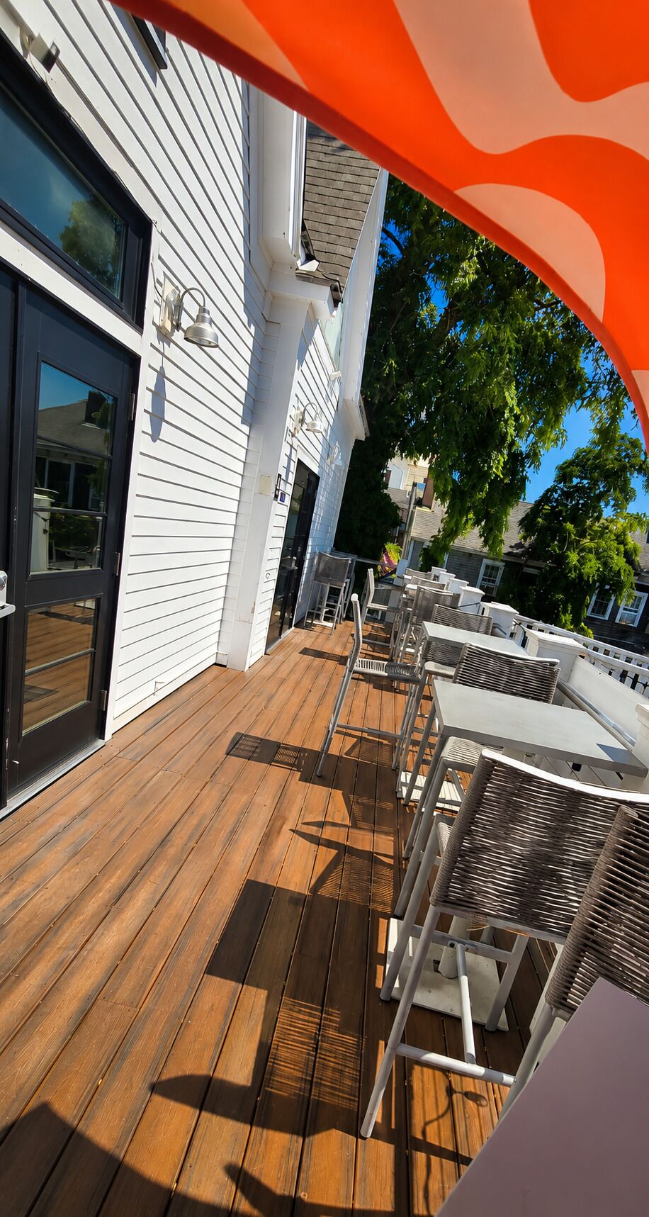 Una terraza de madera con mobiliario exterior, un edificio blanco con una puerta negra y un árbol al fondo.