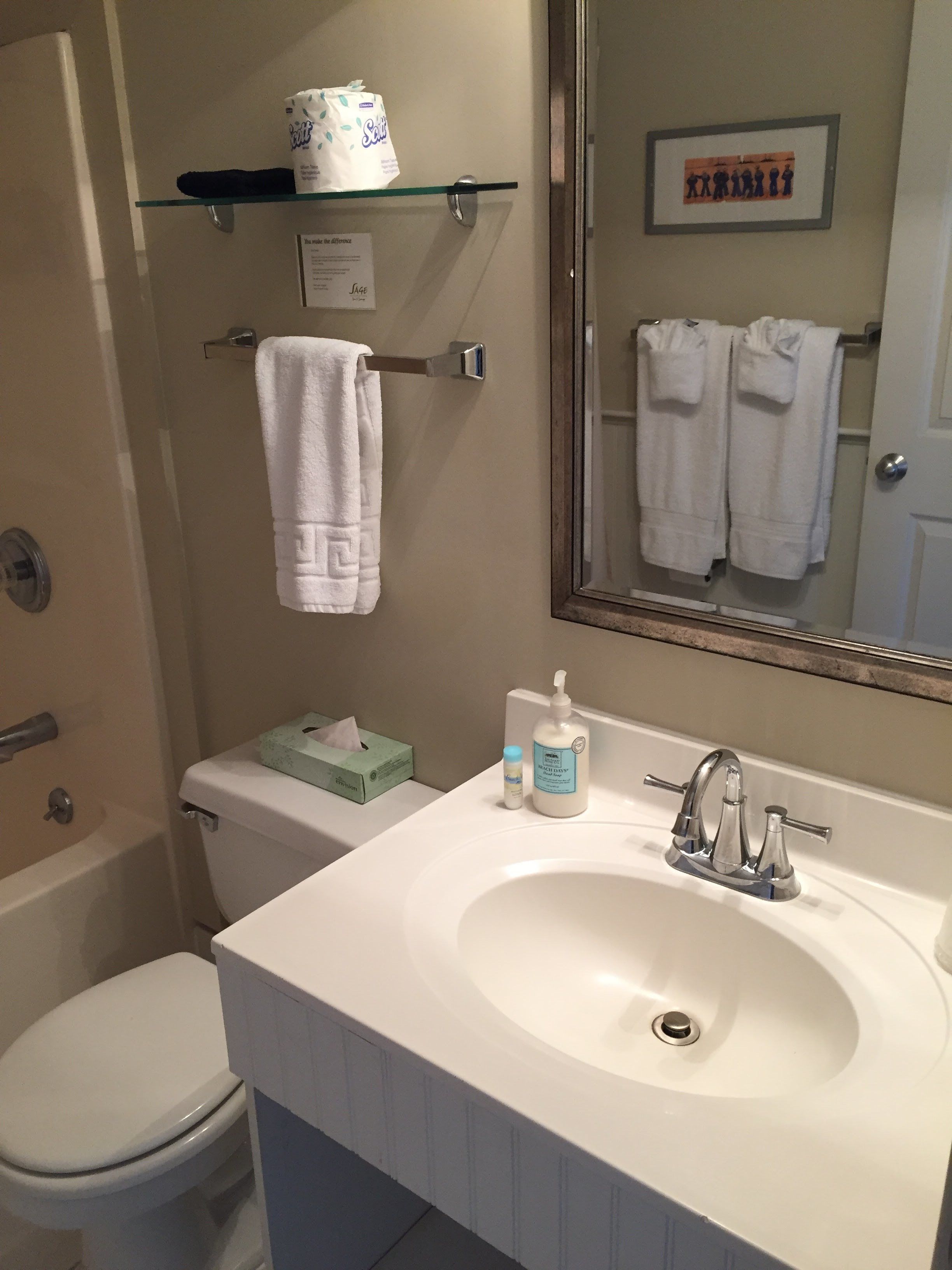A bathroom with a white sink, a mirror, a toilet, and a shower area. There are towels hanging on a rack and a box of tissues on the shelf.