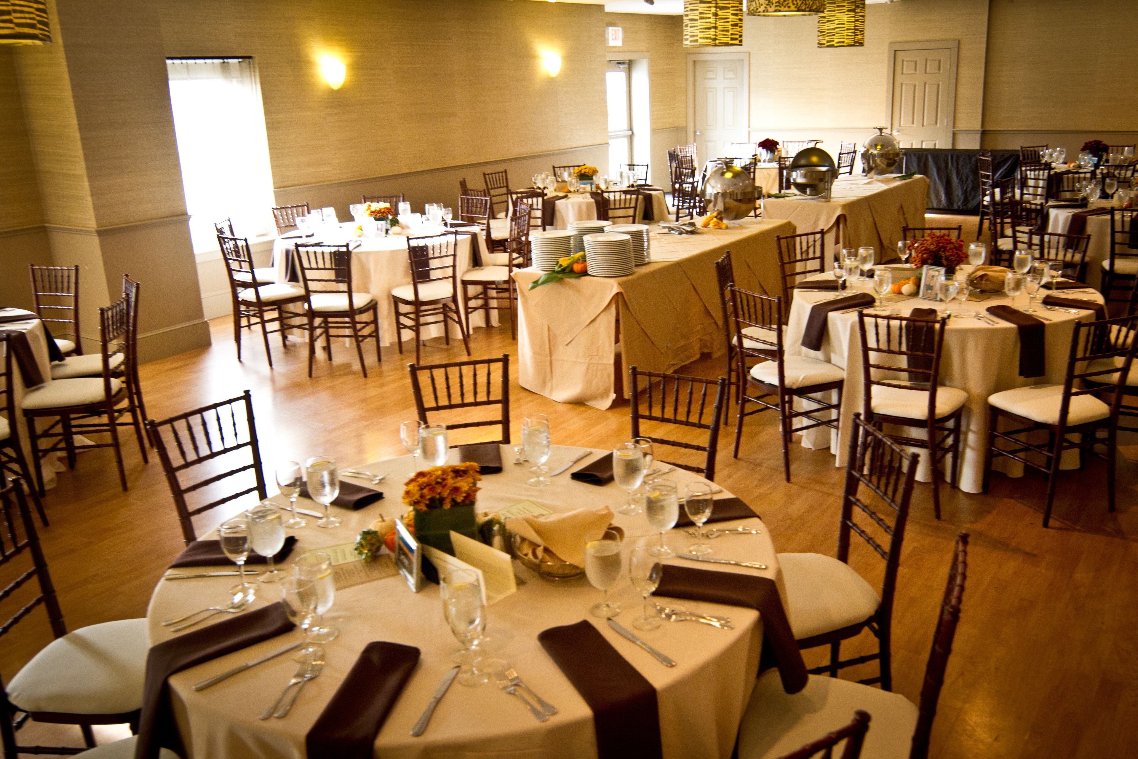 Uma sala de banquetes com várias mesas redondas preparadas para refeições, cada uma com cadeiras e louças.