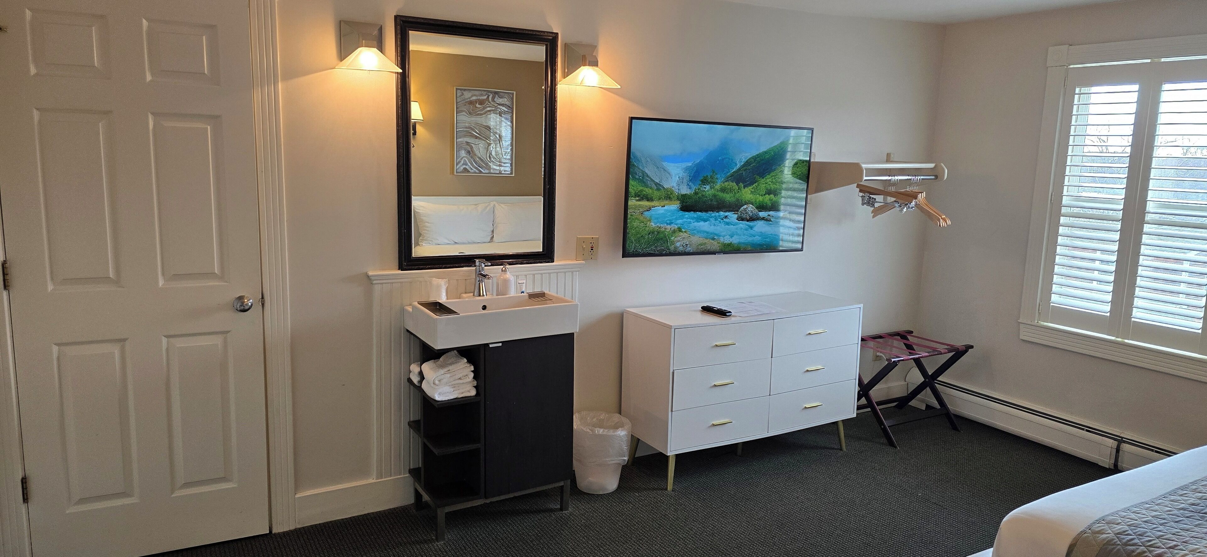 A hotel room with a white door, a sink with a mirror, a white dresser, a painting of a river, and a window with blinds.