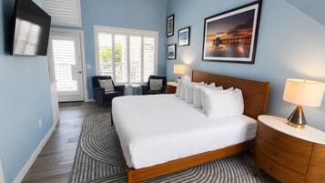 A hotel room with a large bed, a wooden nightstand, a desk, and a chair. There are two windows with blinds, a TV mounted on the wall, and framed pictures on the wall.
