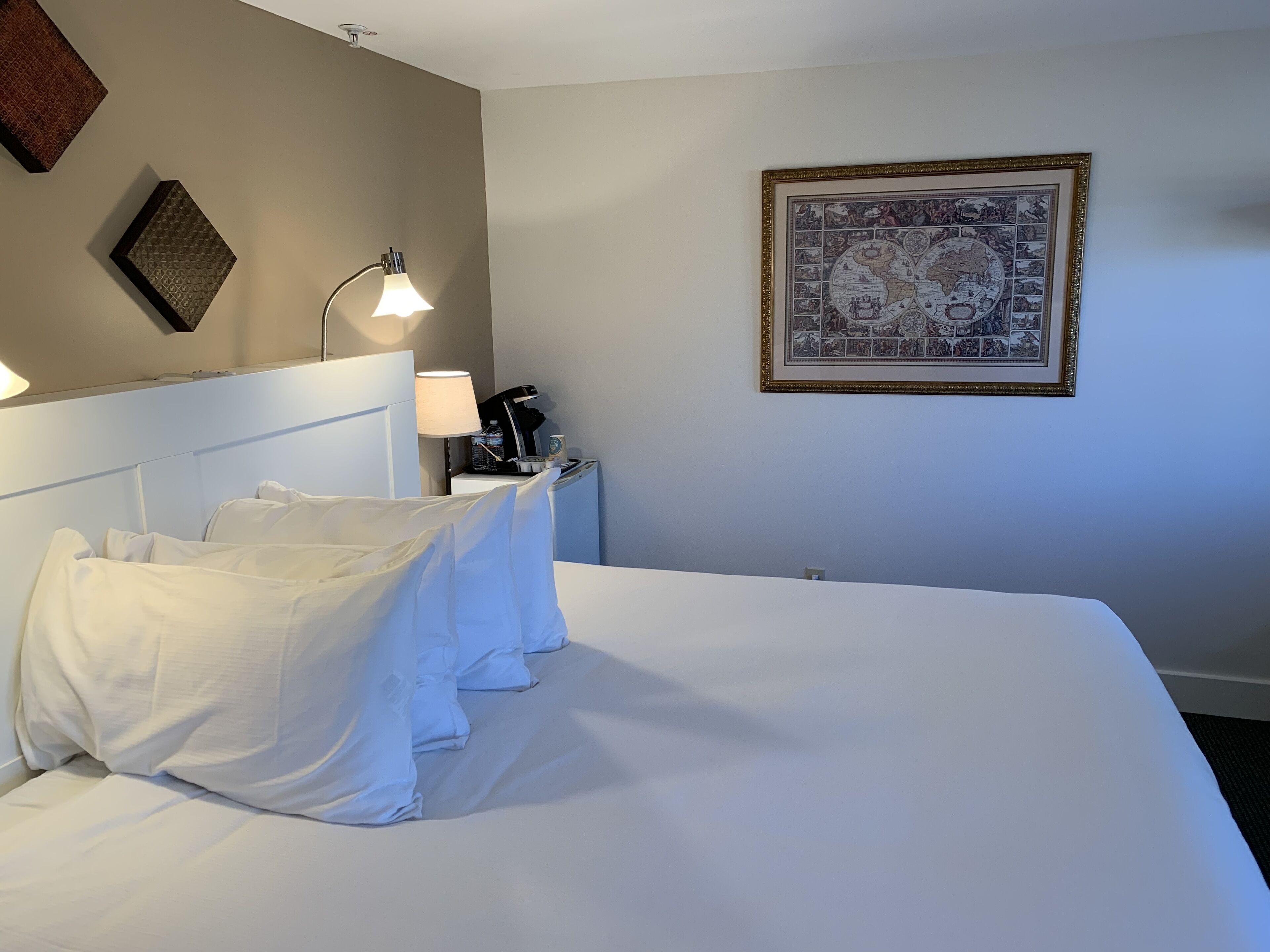 Una habitación de hotel con una cama grande, una mesita de noche con lámpara, un cuadro en la pared y una pequeña mesa con una cafetera.