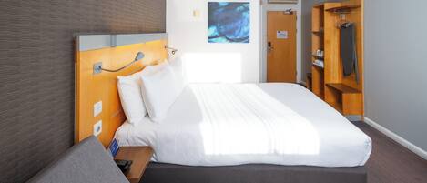 A modern hotel room with a large bed, a wooden headboard, a bedside table, a wardrobe, and a wall-mounted artwork.
