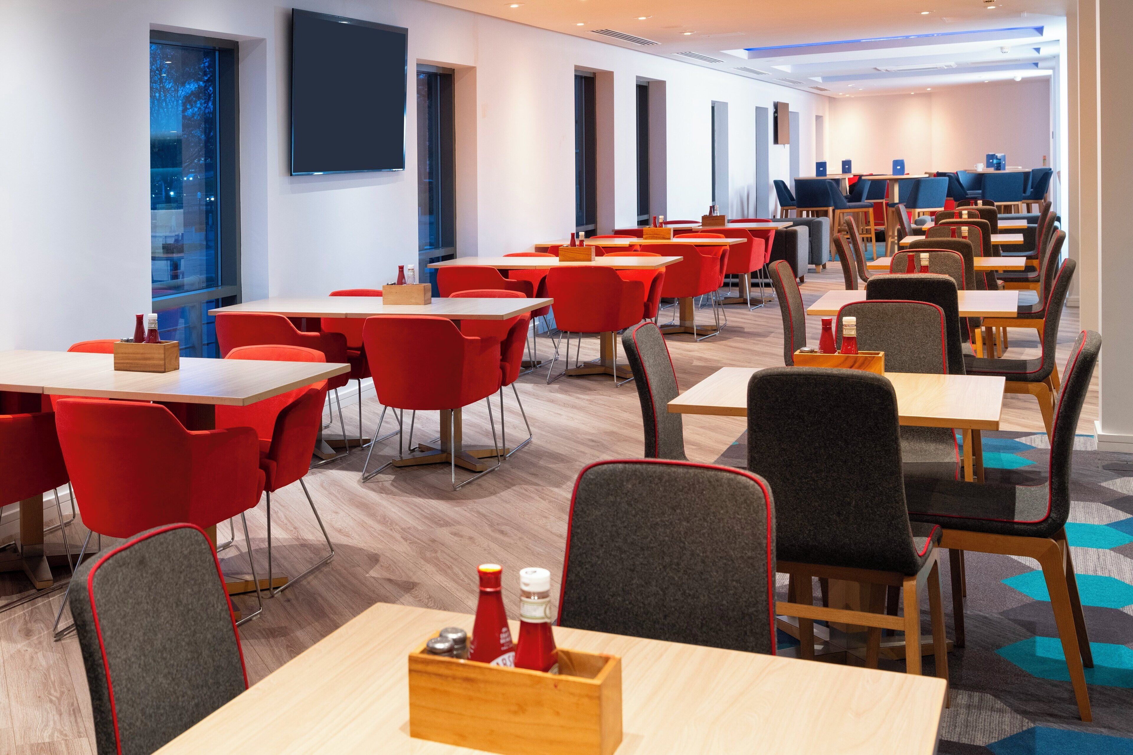 A modern dining area with red and grey chairs, wooden tables, and a blue carpet.