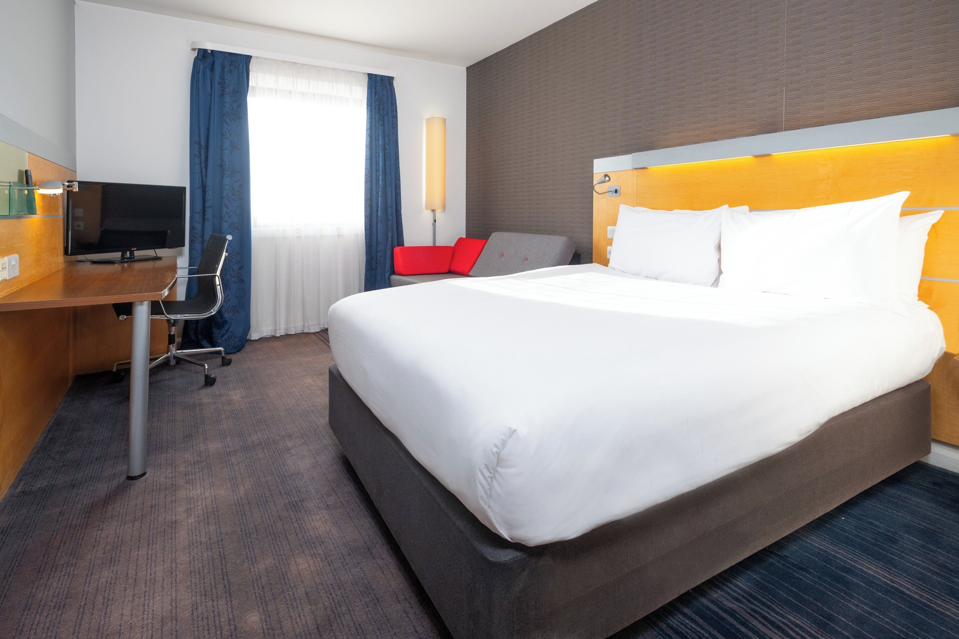 A hotel room with a large bed, a desk with a TV, a chair, and a red accent chair.