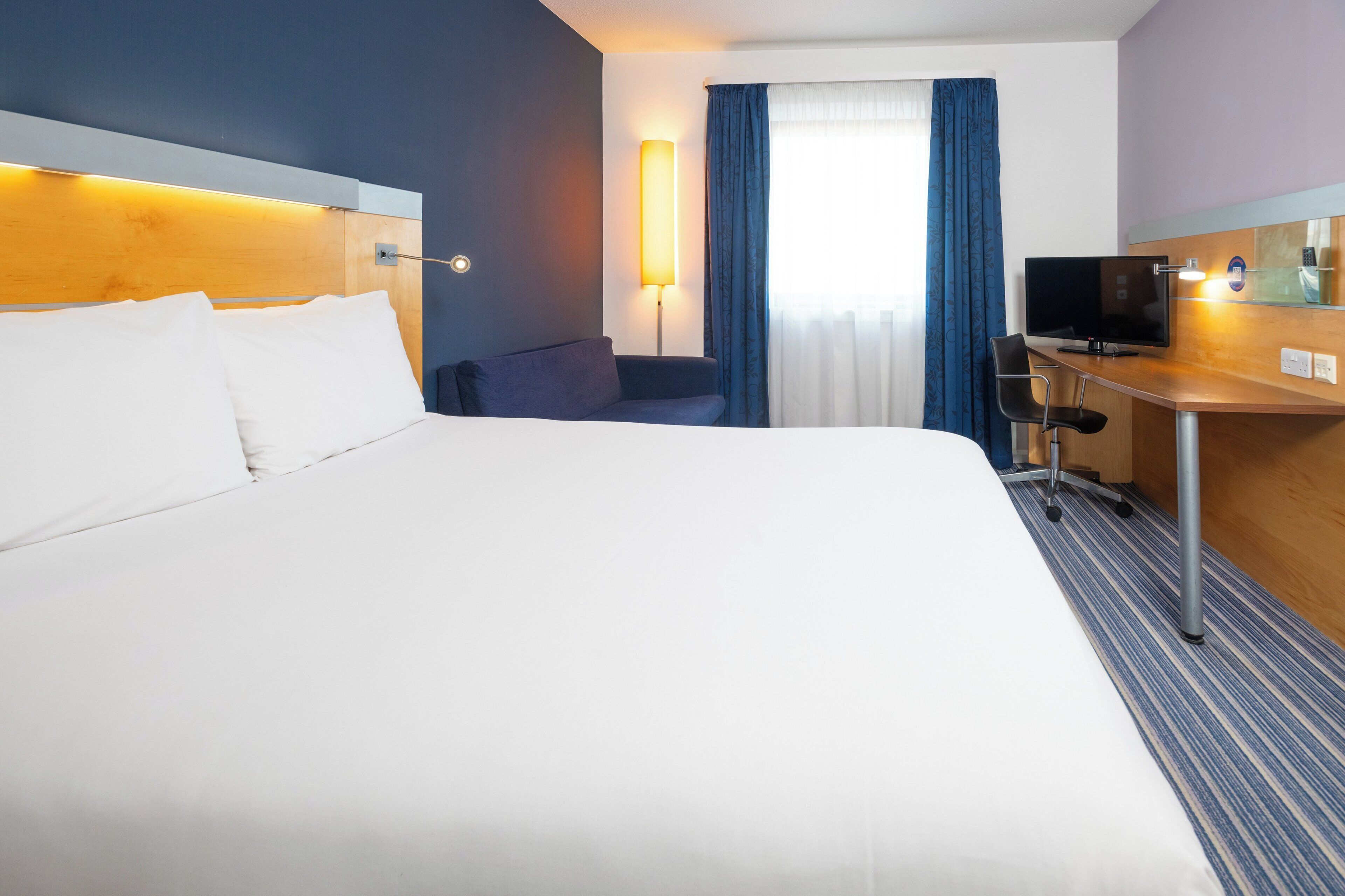 Habitación de hotel con una cama grande, un escritorio con televisor, una lámpara y una ventana con cortinas.