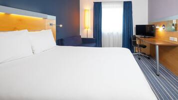 Habitación de hotel con una cama grande, un escritorio con televisor, una lámpara y una ventana con cortinas.