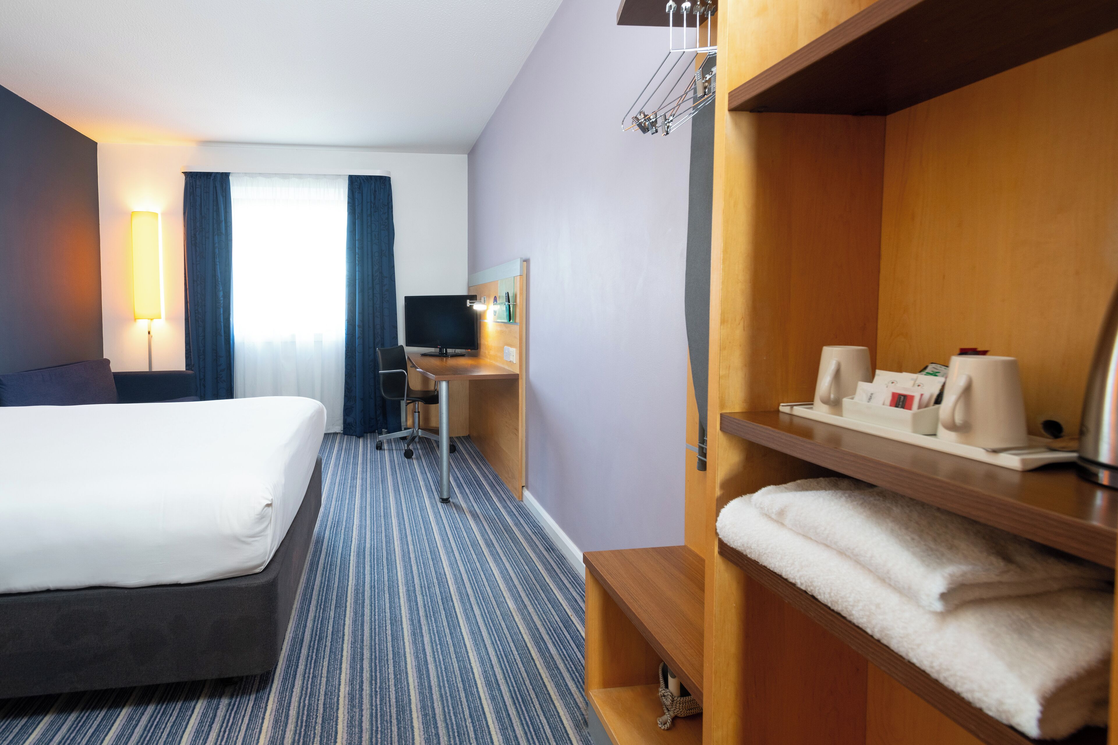 A hotel room with two beds, a desk, a chair, a TV, a lamp, and a shelf with towels and toiletries.