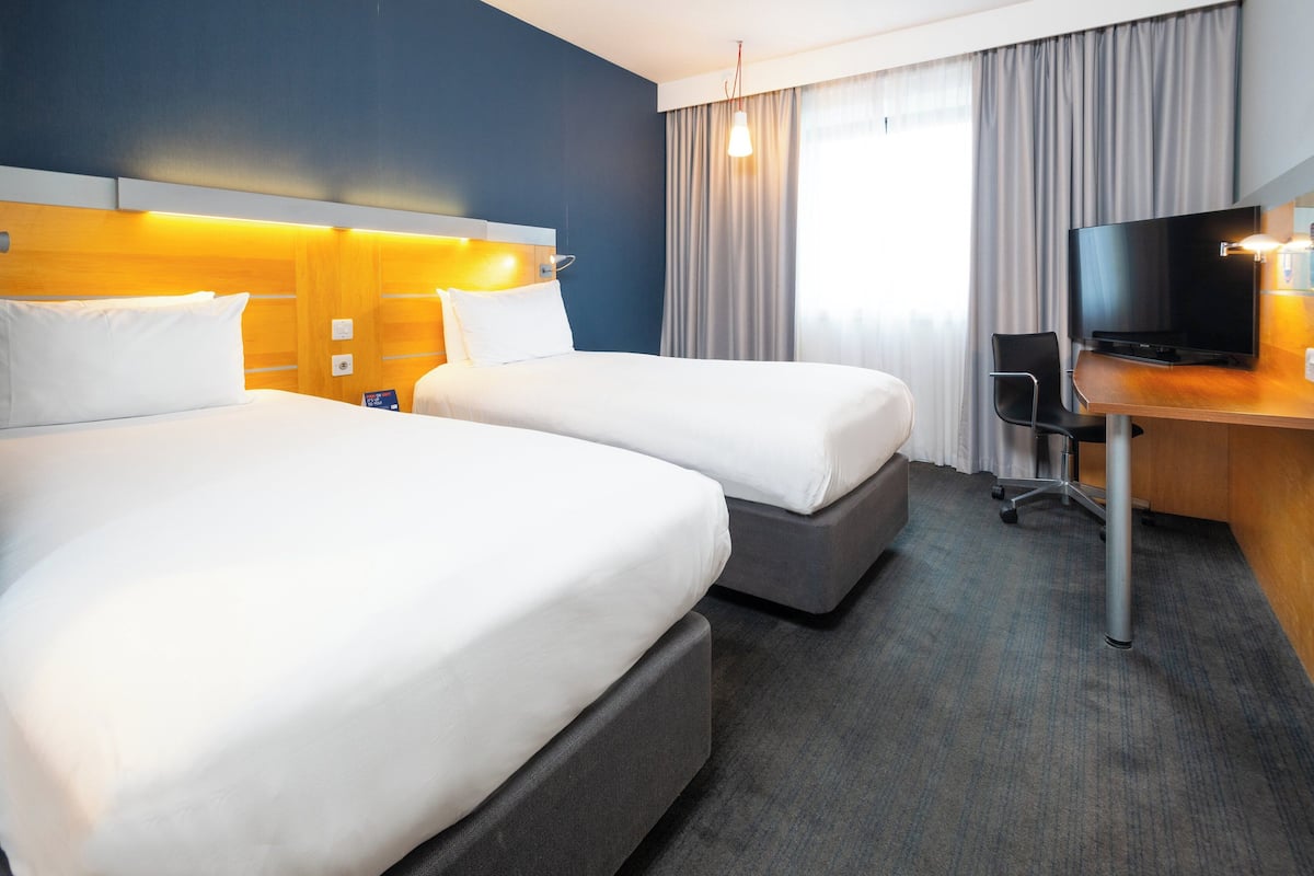 A hotel room with two beds, a desk with a television, and a large window with curtains.