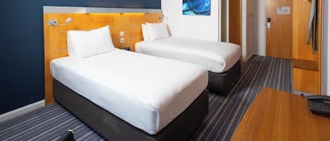 A hotel room with two beds, a desk, a chair, a wooden headboard, a blue abstract painting, and a door with a towel rack.
