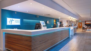 A Holiday Inn Express reception area with a modern design, featuring a blue accent wall, a wooden reception desk, and a seating area with tables and chairs. - Holiday Inn Express Stevenage by IHG (Stevenage)