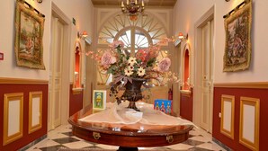 A grand hallway with a large wooden table at the center, surrounded by red walls and gold trim, leading to a grand arched doorway. - Halepa Hotel (Chania)