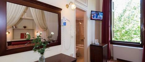 A hotel room with a wooden desk, a vase with flowers, a mirror, a television, a shower, and a window with a view of greenery.