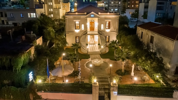 A large, historic building with a central staircase, surrounded by a well-maintained garden and outdoor seating area. - Halepa Hotel (Chania)