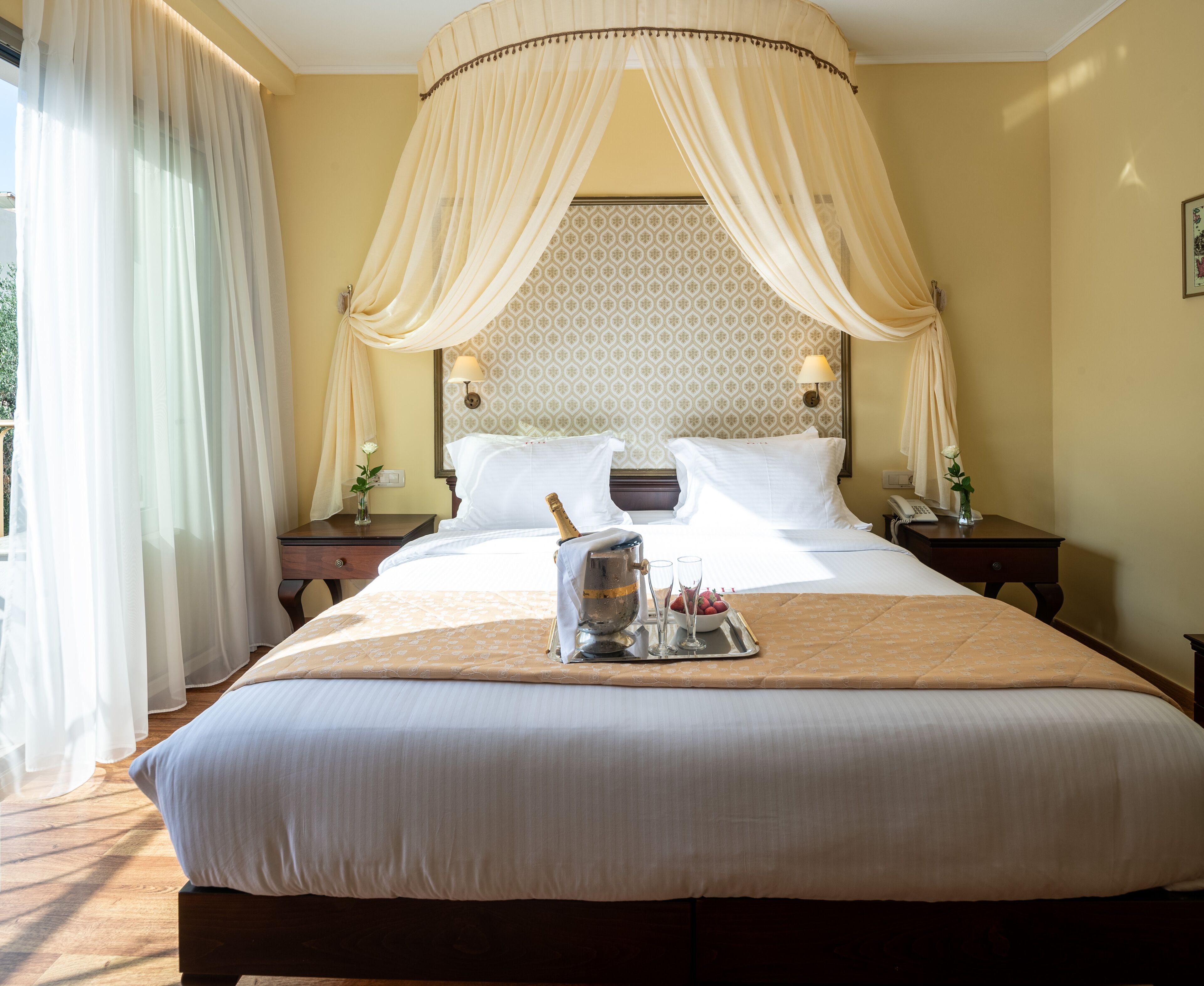 A large bed with a canopy, a bedside table with a lamp, and a tray with a bottle and glasses.