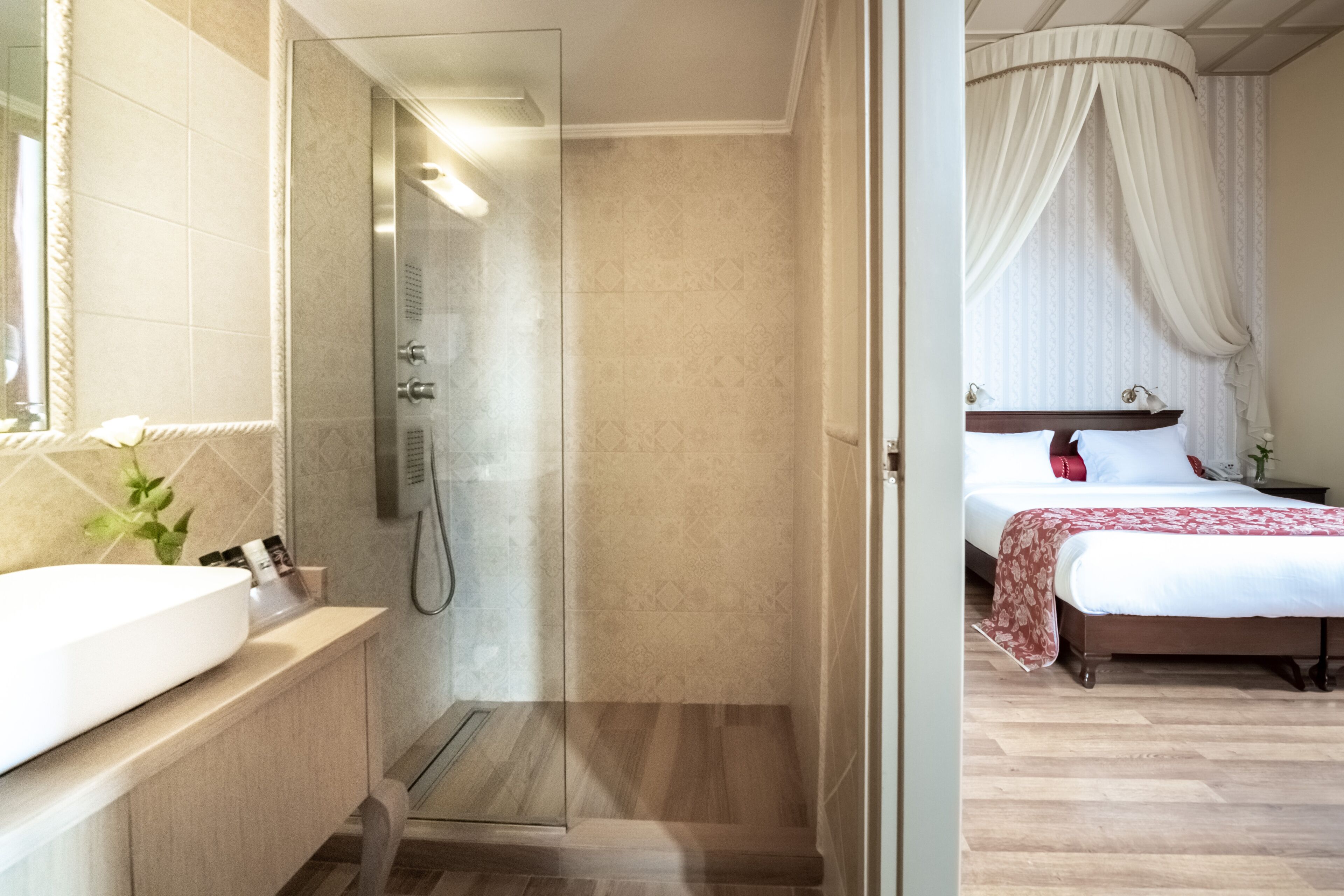 A bathroom with a glass-enclosed shower, a sink with a mirror, and a bedside table with a lamp.
