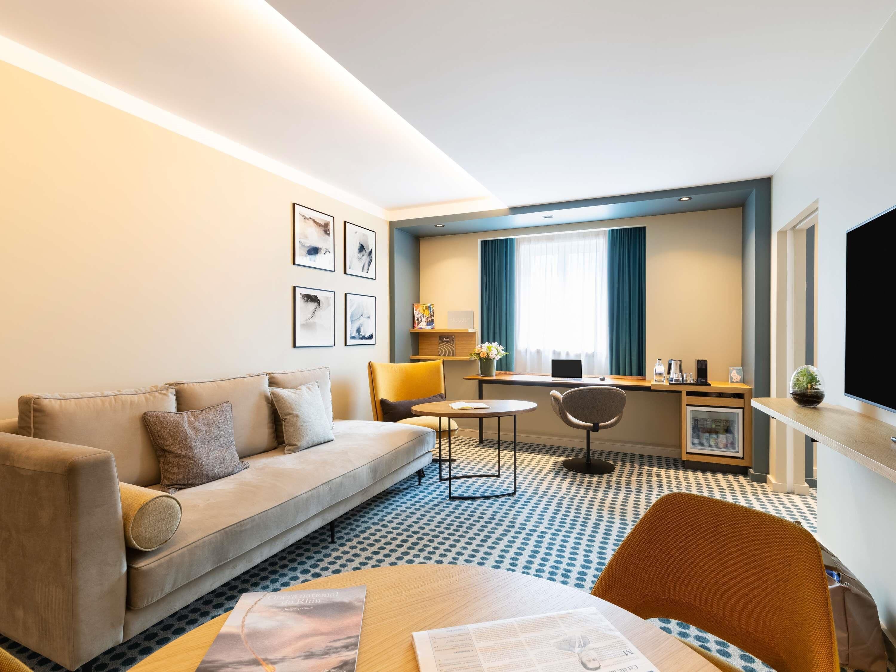 Habitación de hotel con sofá, mesa de comedor, computadora portátil, televisión y obras de arte en la pared.
