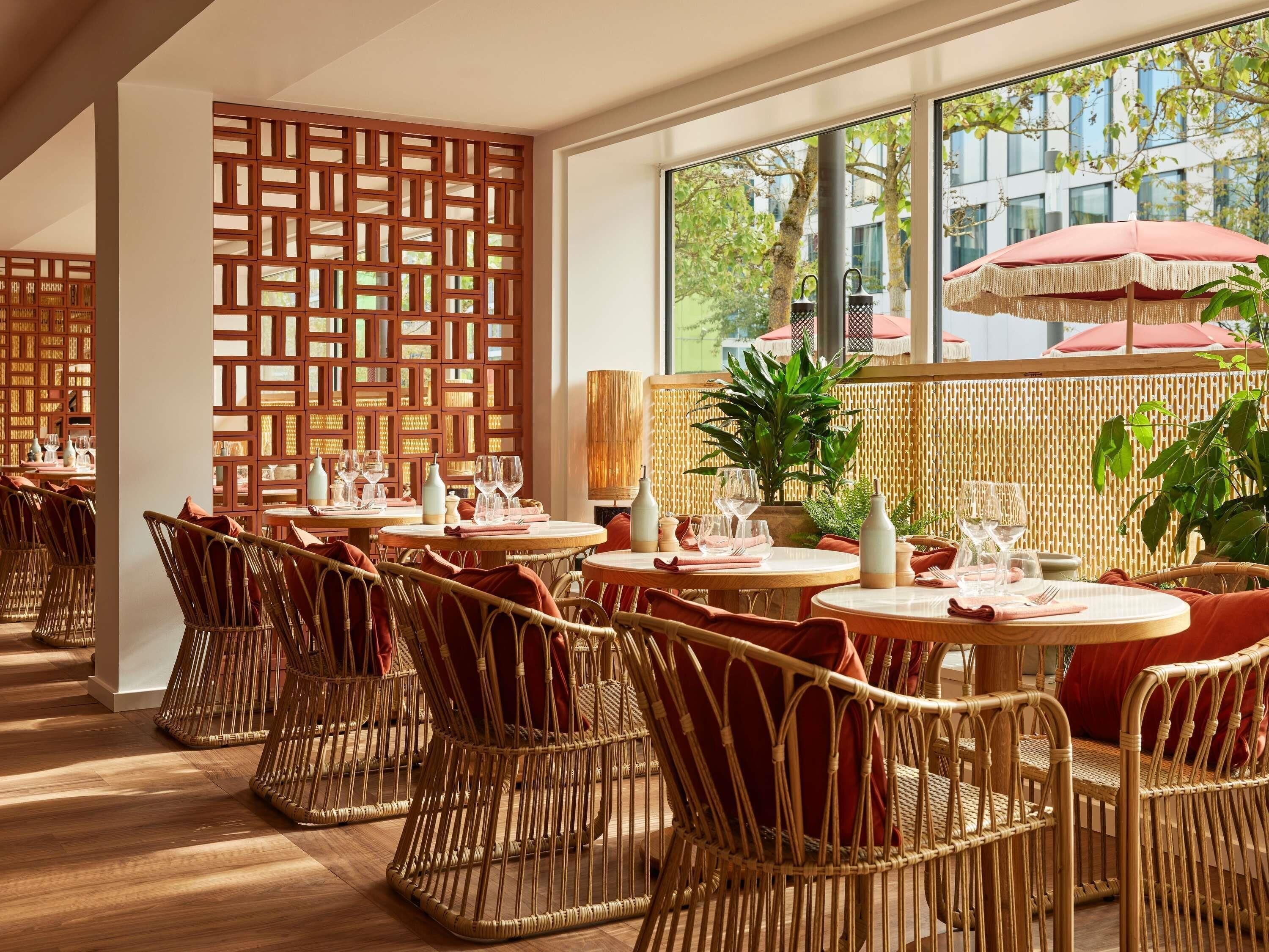 Un restaurant avec des chaises en osier, des tables rondes et une vue sur l’extérieur grâce à de grandes fenêtres.